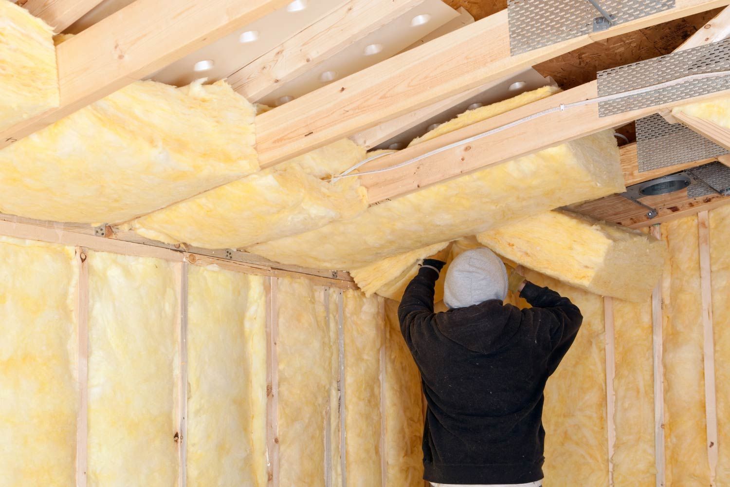 Person installing batt insulation in a wood-framed wall and ceiling. Yellow insulation fills spaces.