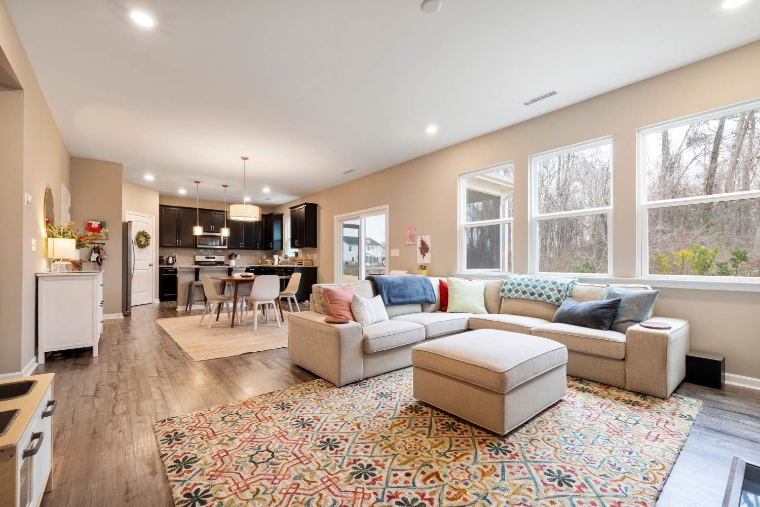 Living room with beige sectional sofa, patterned rug, open to kitchen and large windows overlooking trees.