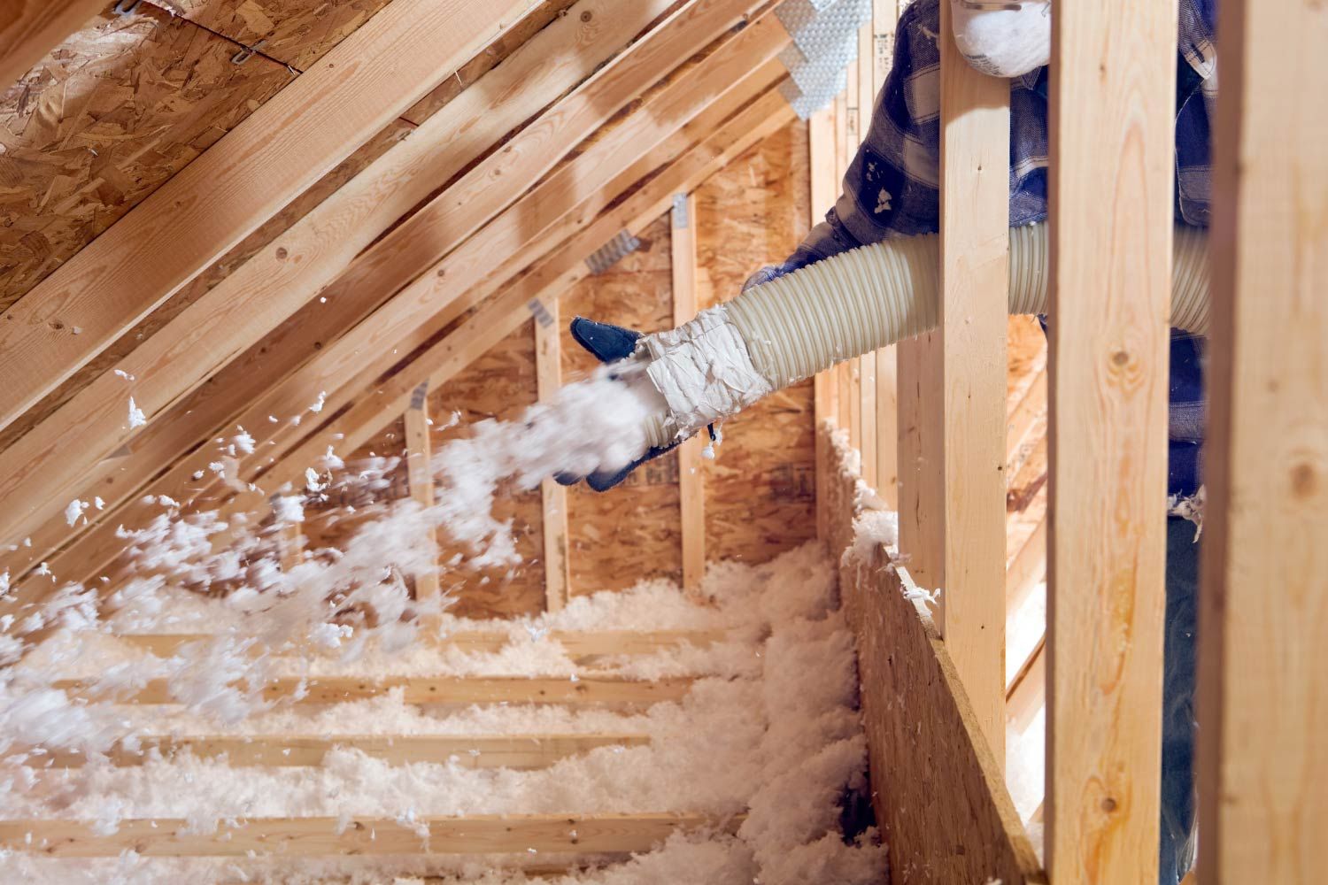 Person blowing insulation into an attic. Wooden framing and white insulation are visible.