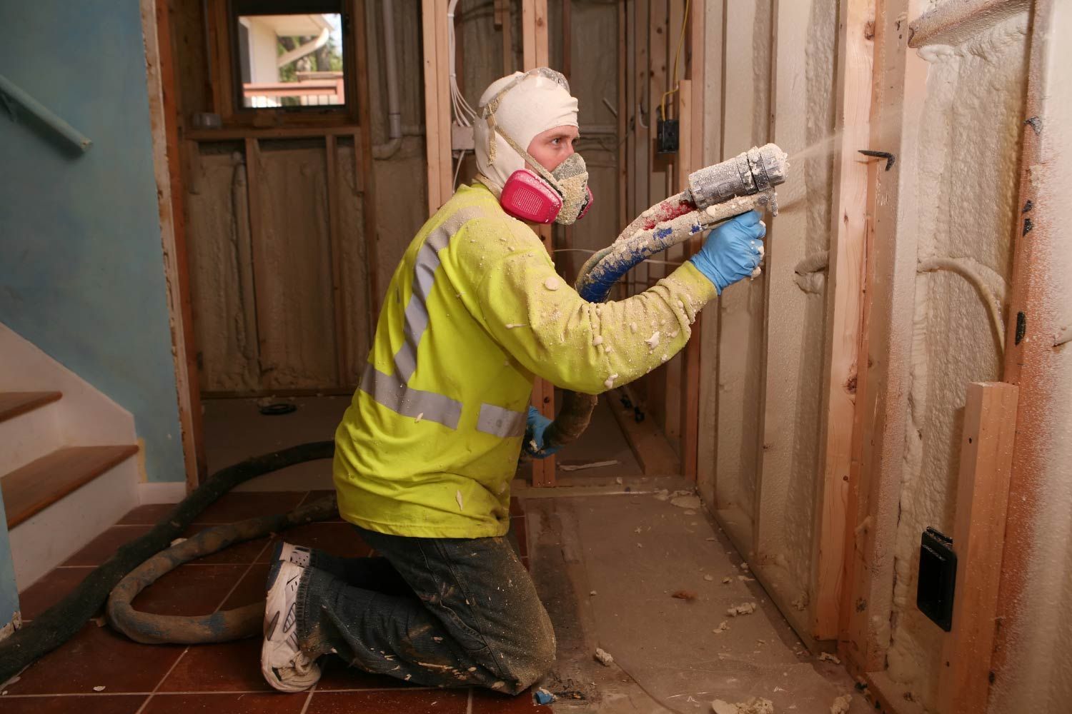 Man in protective gear sprays insulation into a wall. Yellow jacket, respirator, kneeling indoors.