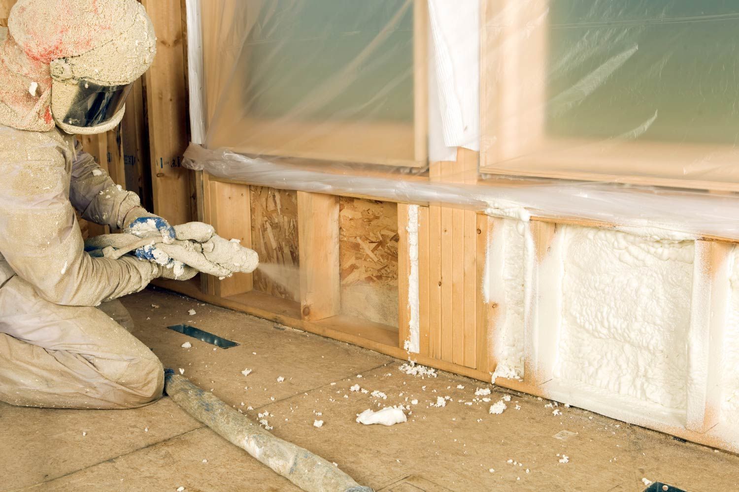 Person kneeling, spraying foam insulation into a wooden wall frame; safety gear worn.