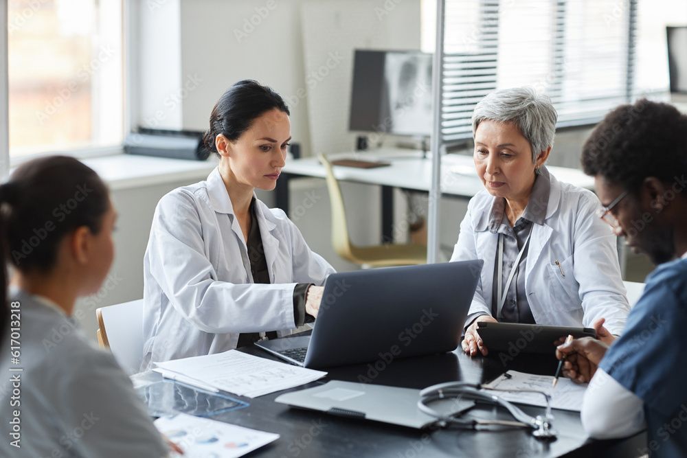 A group of doctors are sitting around a table looking at a laptop.