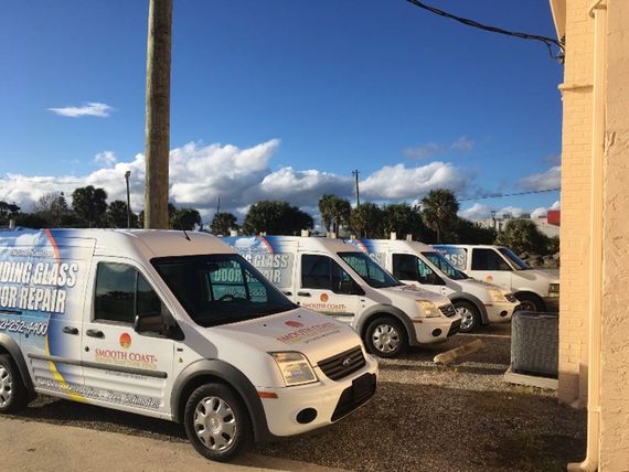 White service vans with logos parked in a row, under a bright blue sky. The vans are likely for glass repair.