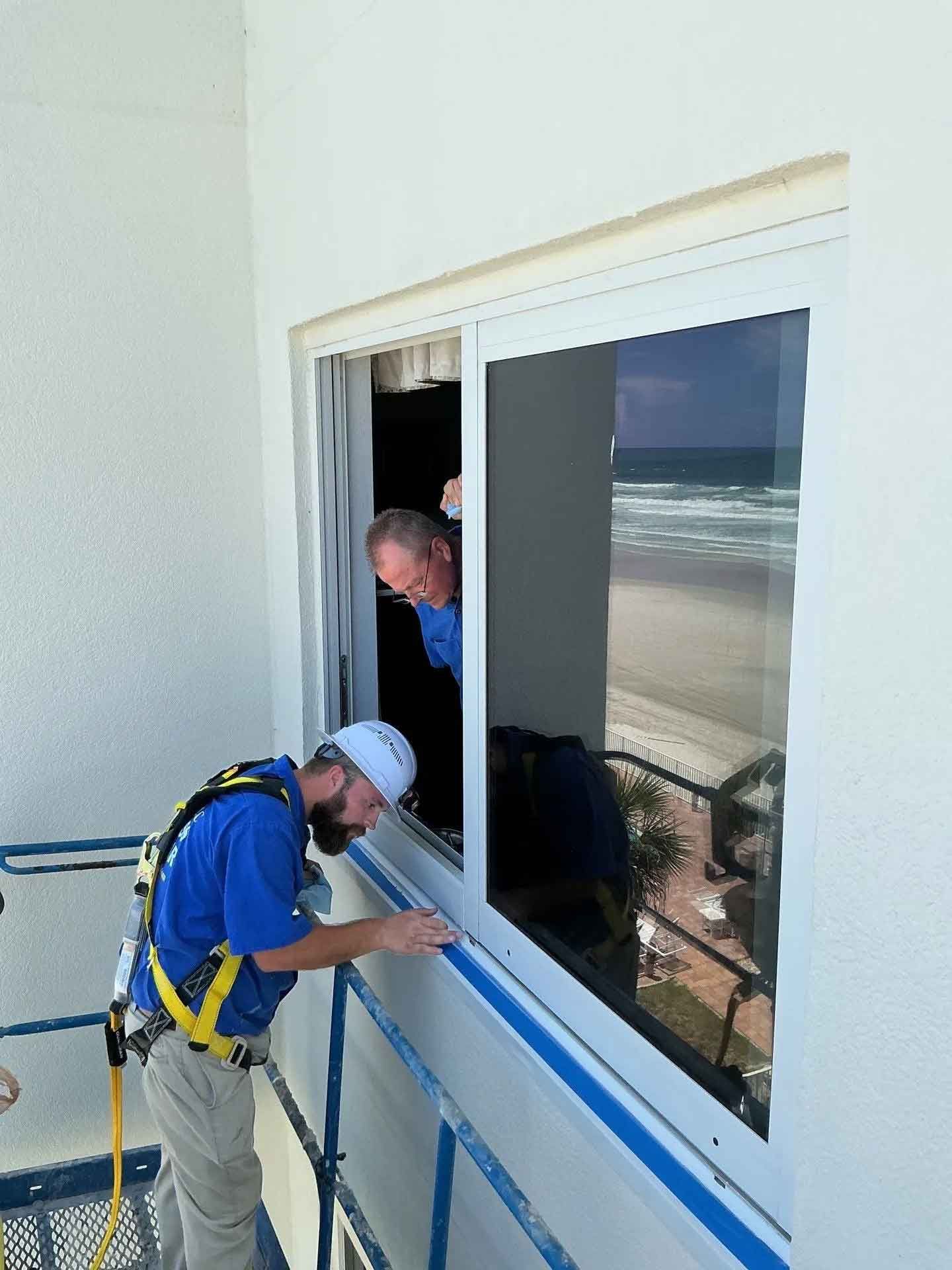 Two workers on a suspended scaffold install a window overlooking a beach.
