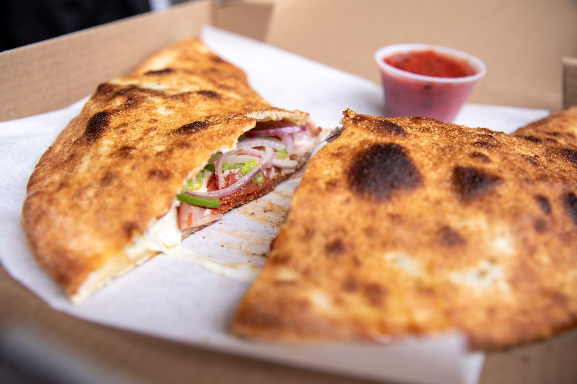 Calzone cut open, revealing filling of vegetables and cheese, served with red dipping sauce.