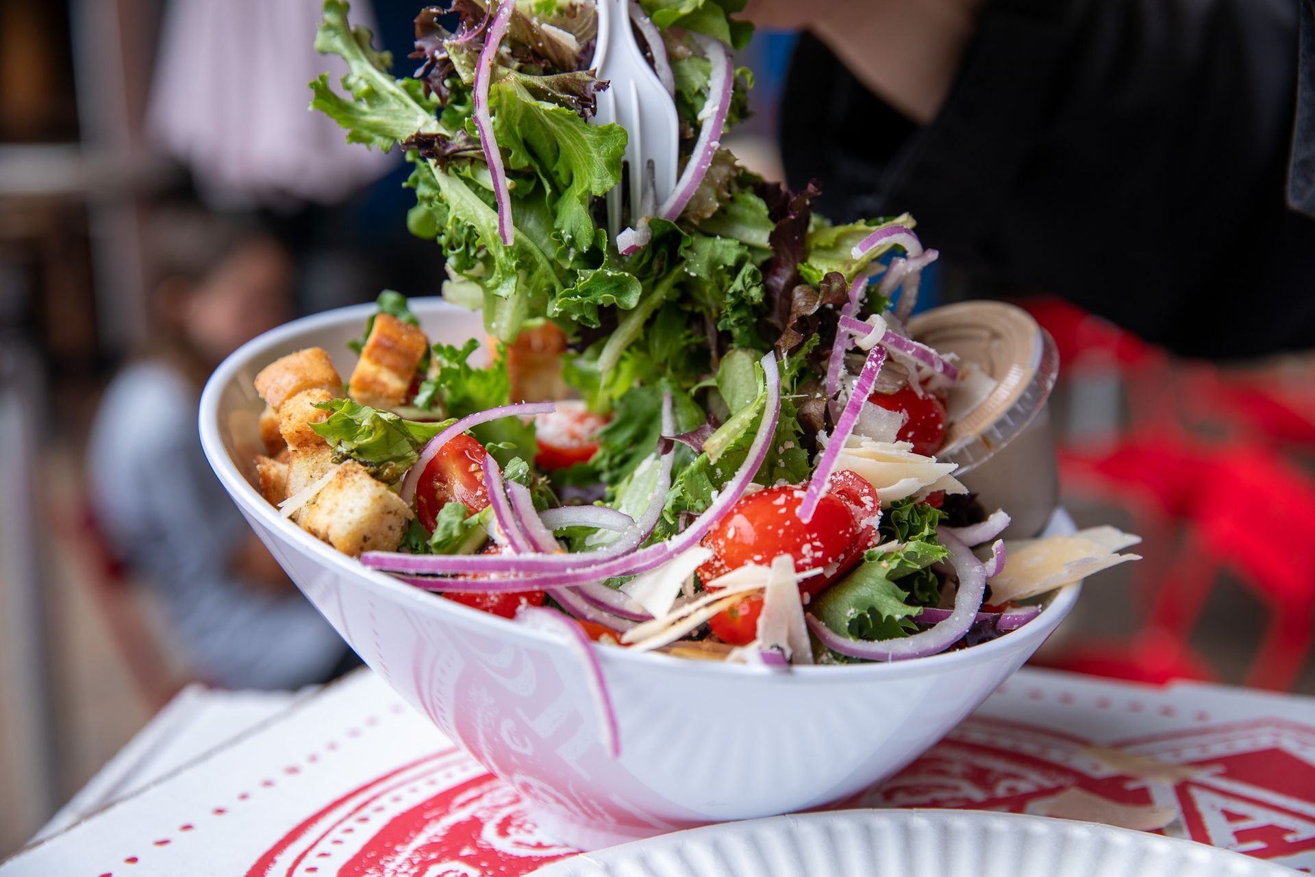 Person tossing salad with red onion, tomatoes, croutons, and parmesan cheese in a white bowl.