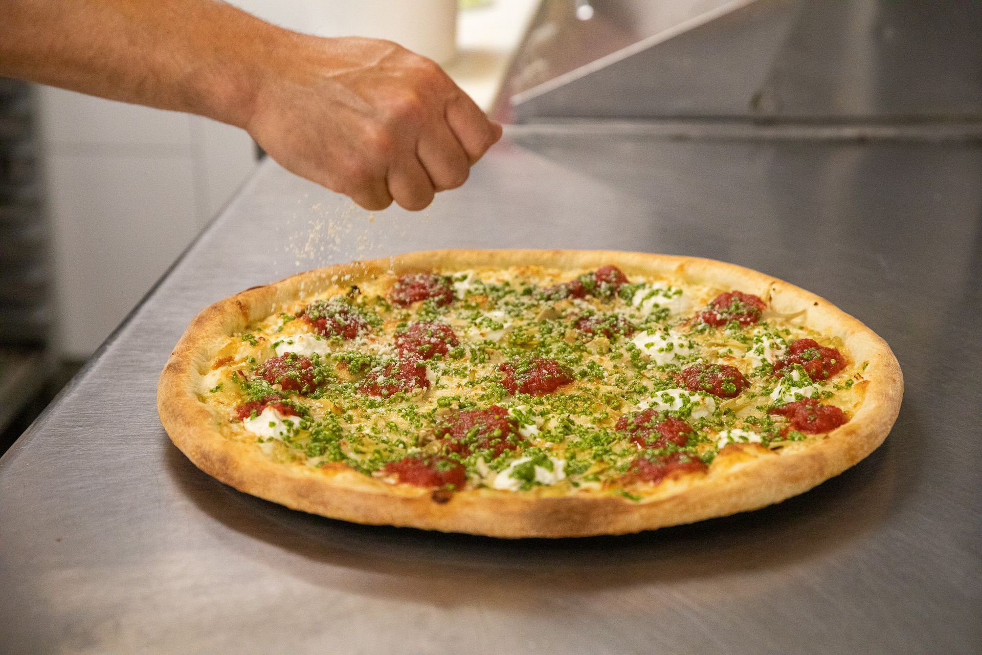A hand sprinkling topping on a finished pizza with red and green ingredients, on a stainless steel surface.