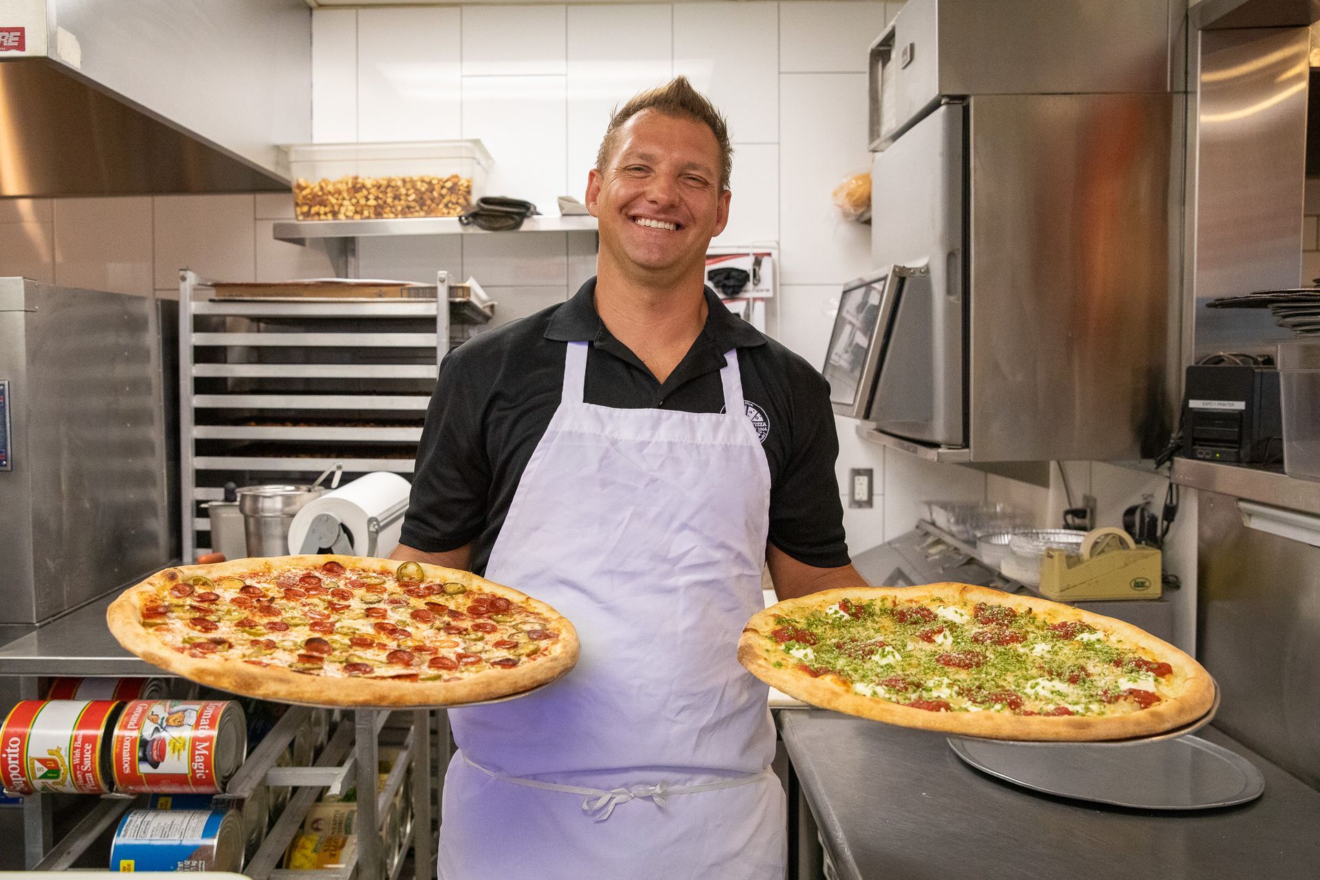 Man in apron holding two pizzas, smiling in a commercial kitchen.