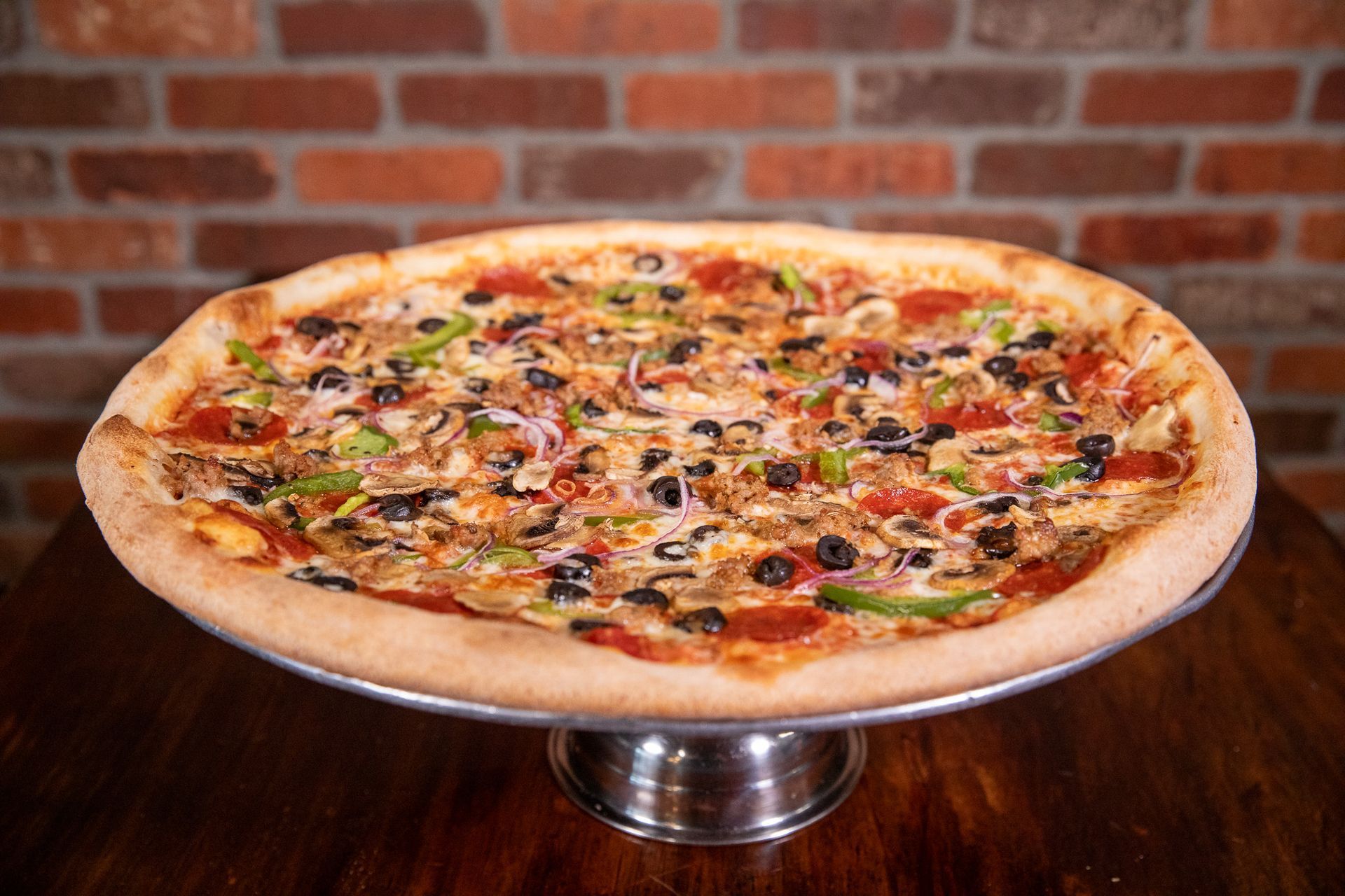 Pizza with various toppings on a silver stand in front of a brick wall.