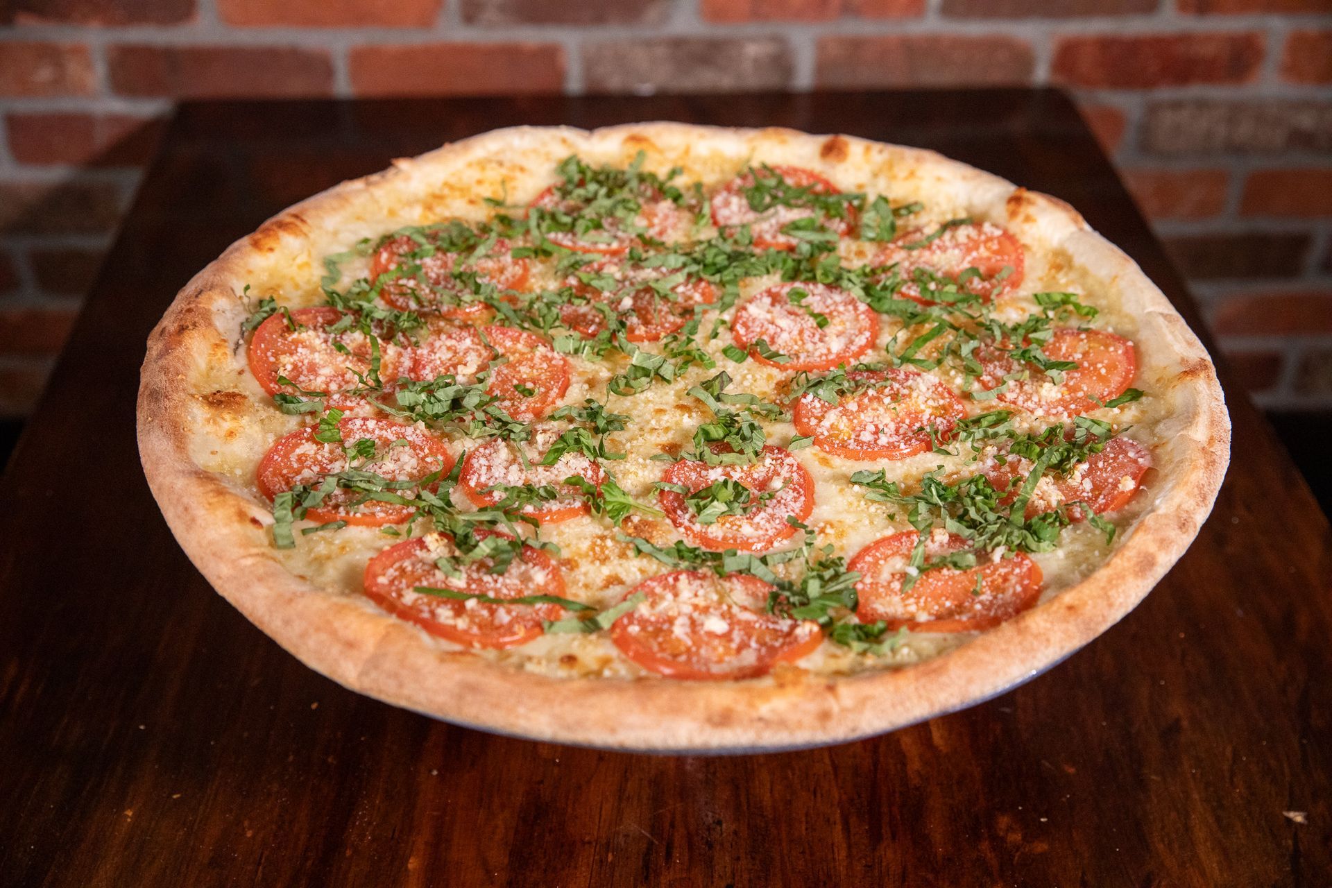 Pizza with tomato slices, herbs, and cheese on a wooden table, against a brick wall backdrop.