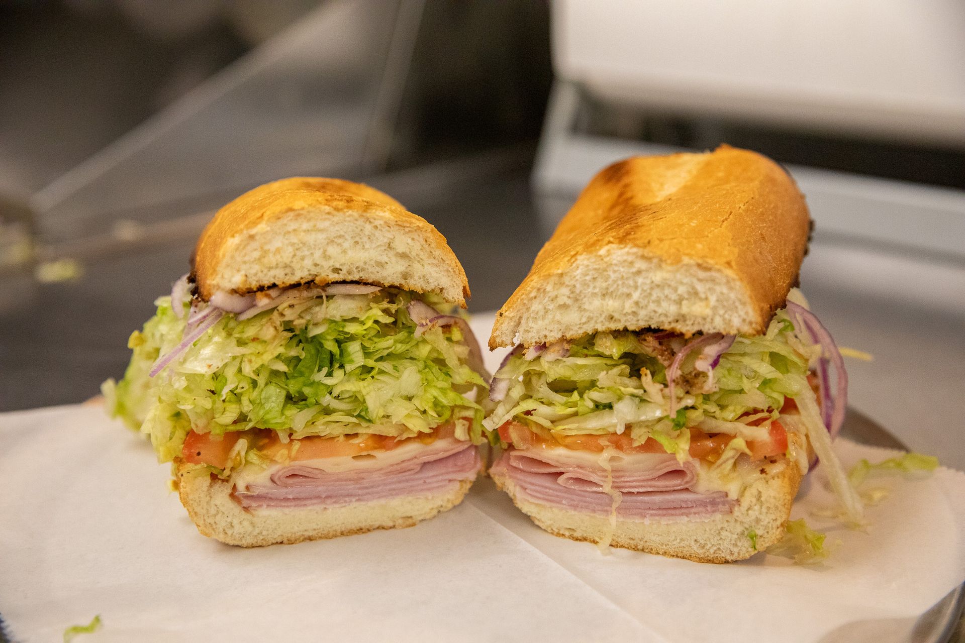 A sub sandwich cut in half, revealing layers of ham, cheese, lettuce, tomatoes, and red onions on a white paper.