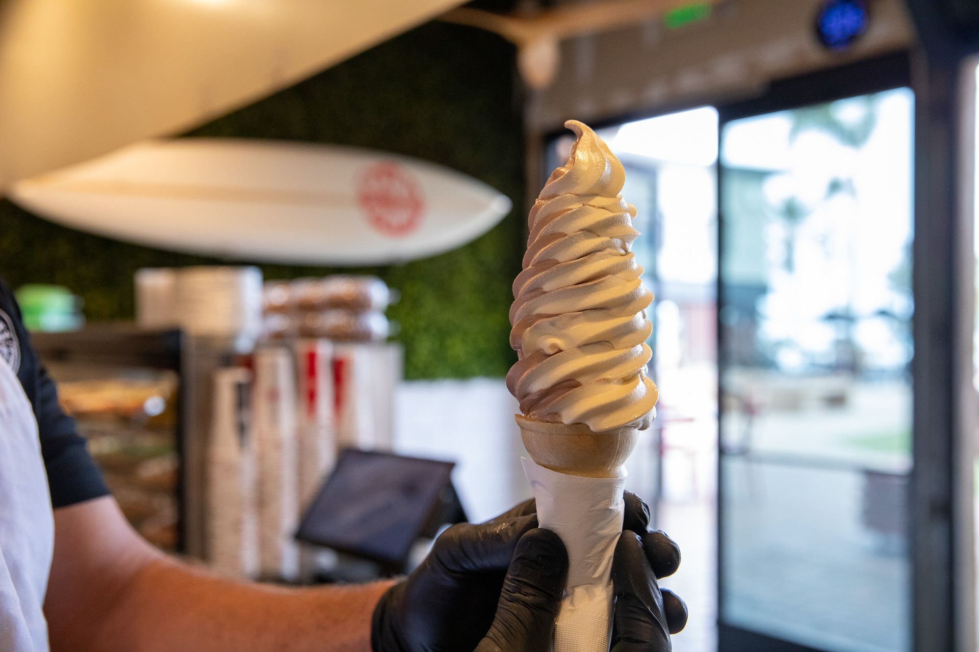 Soft-serve ice cream cone being held, with a surfboard in the background.