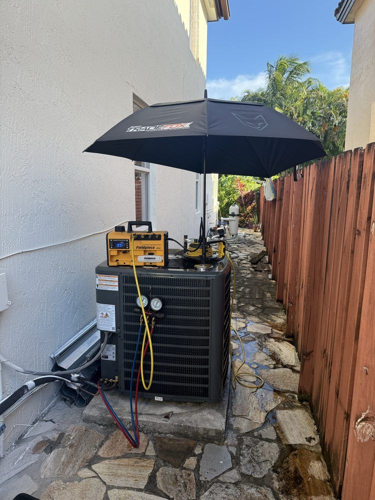 HVAC technician servicing a unit. Yellow gauges, black umbrella, narrow outdoor setting with a fence and stone path.