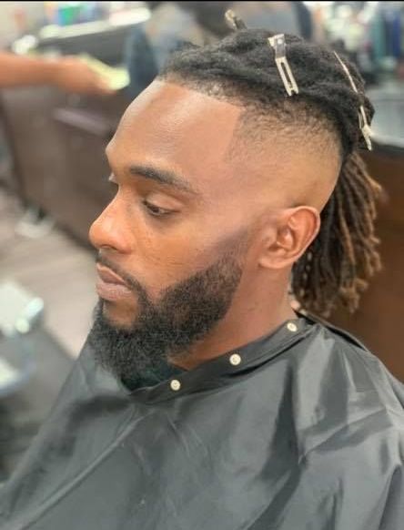 A man with dreadlocks and a beard is getting his hair cut at a barber shop.