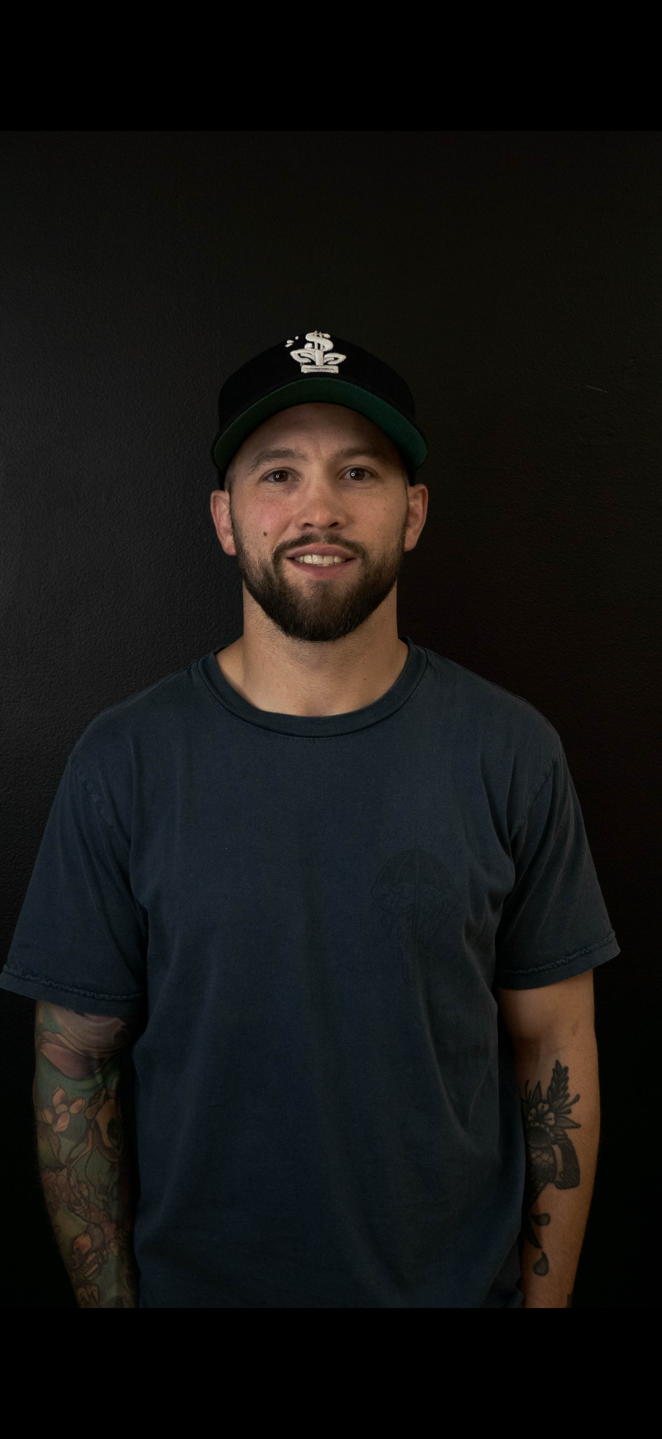 Man wearing a black hoodie and baseball cap smiles at the camera.