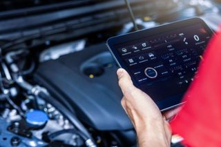 A person in a red shirt uses a digital tablet to inspect a car engine in an auto repair shop.