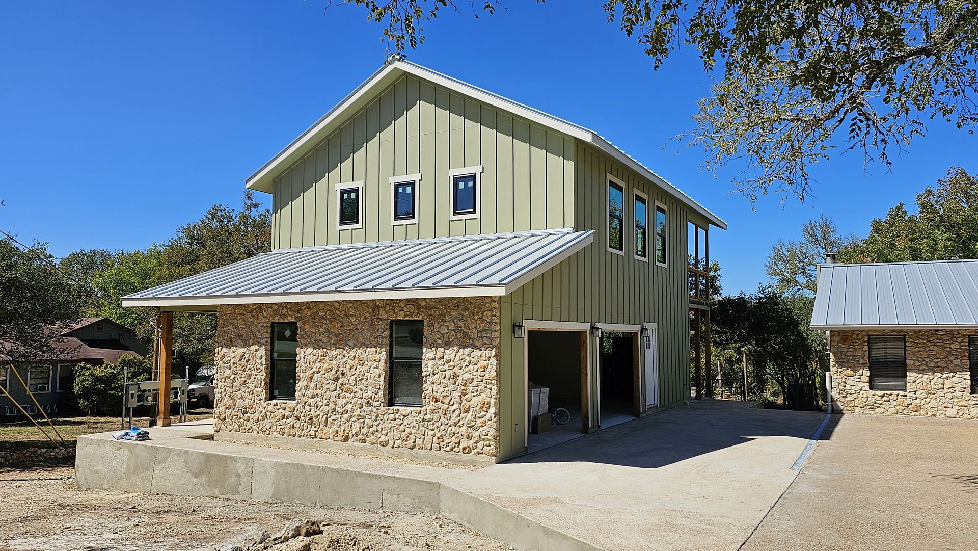 Two-story house with light green siding, stone facade, gray roof, and small adjacent building on a gravel driveway.