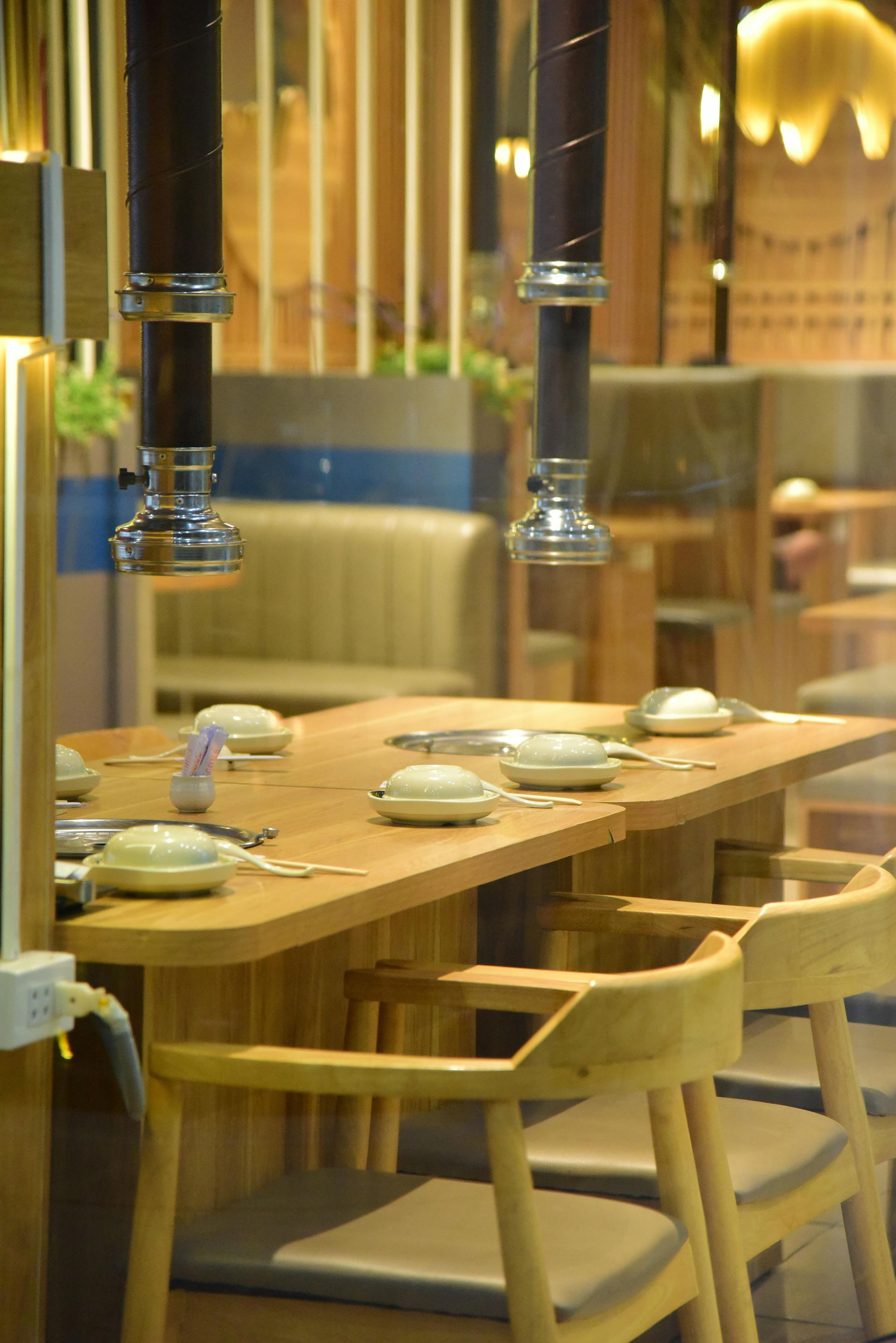 Wooden table with grill and chairs in a restaurant, metal exhaust pipes overhead, natural light.