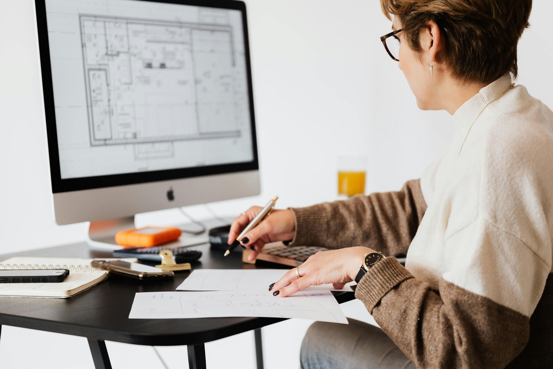 Woman working at desk, reviewing blueprints on computer and paper, with pen in hand.