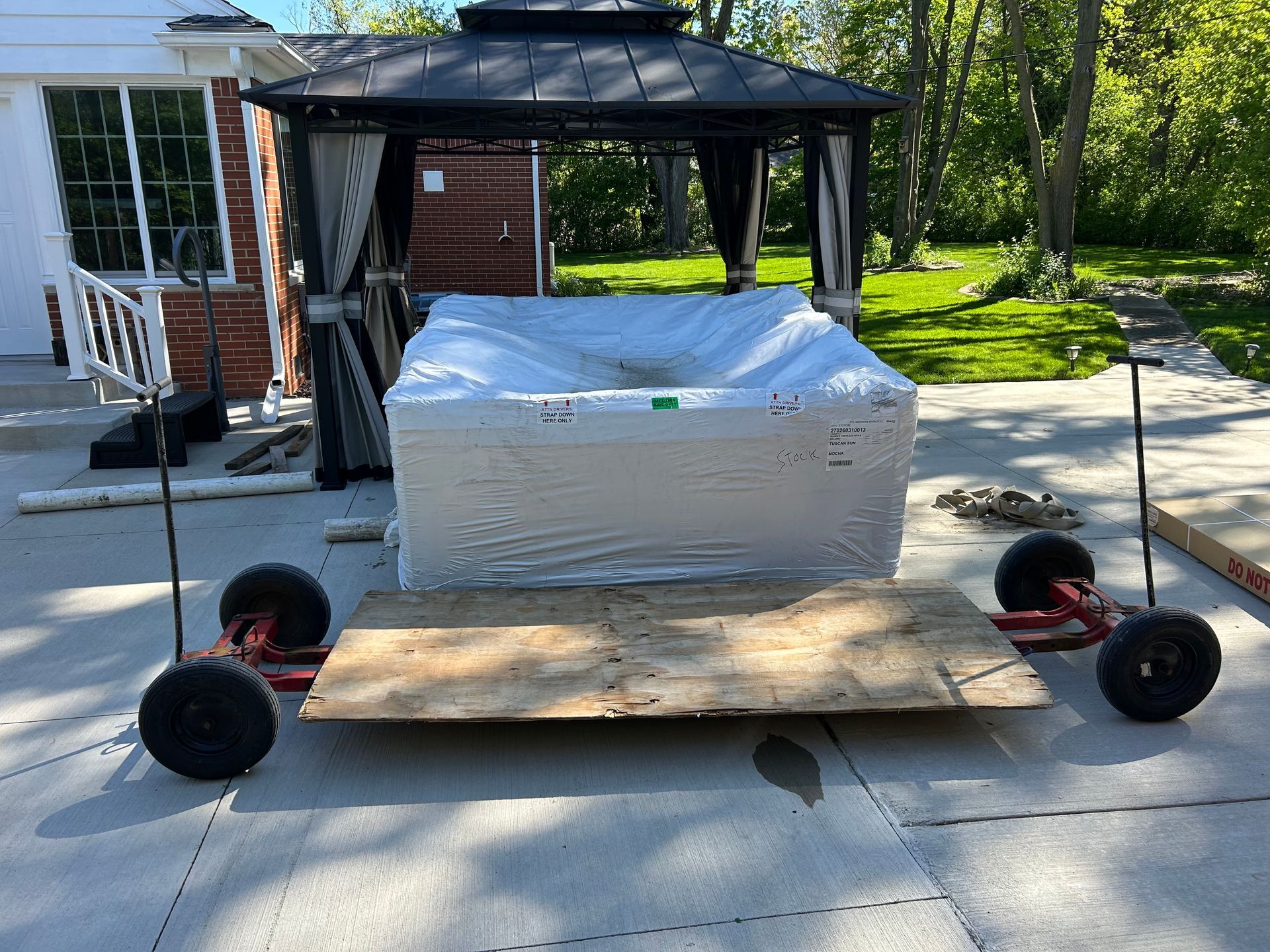 Black-canopy cart on wheels beside a house, with a wrapped white cargo box on its platform.