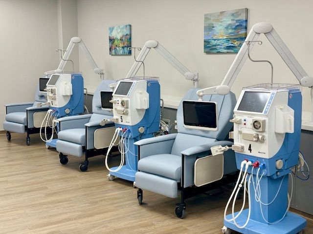 Four blue dialysis machines with connected chairs in a medical facility.