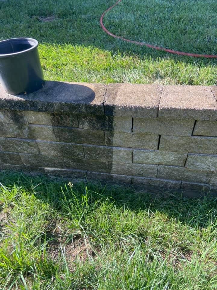 A retaining wall half cleaned with a pressure washer. Dark algae on the uncleaned side; grass surrounds it.