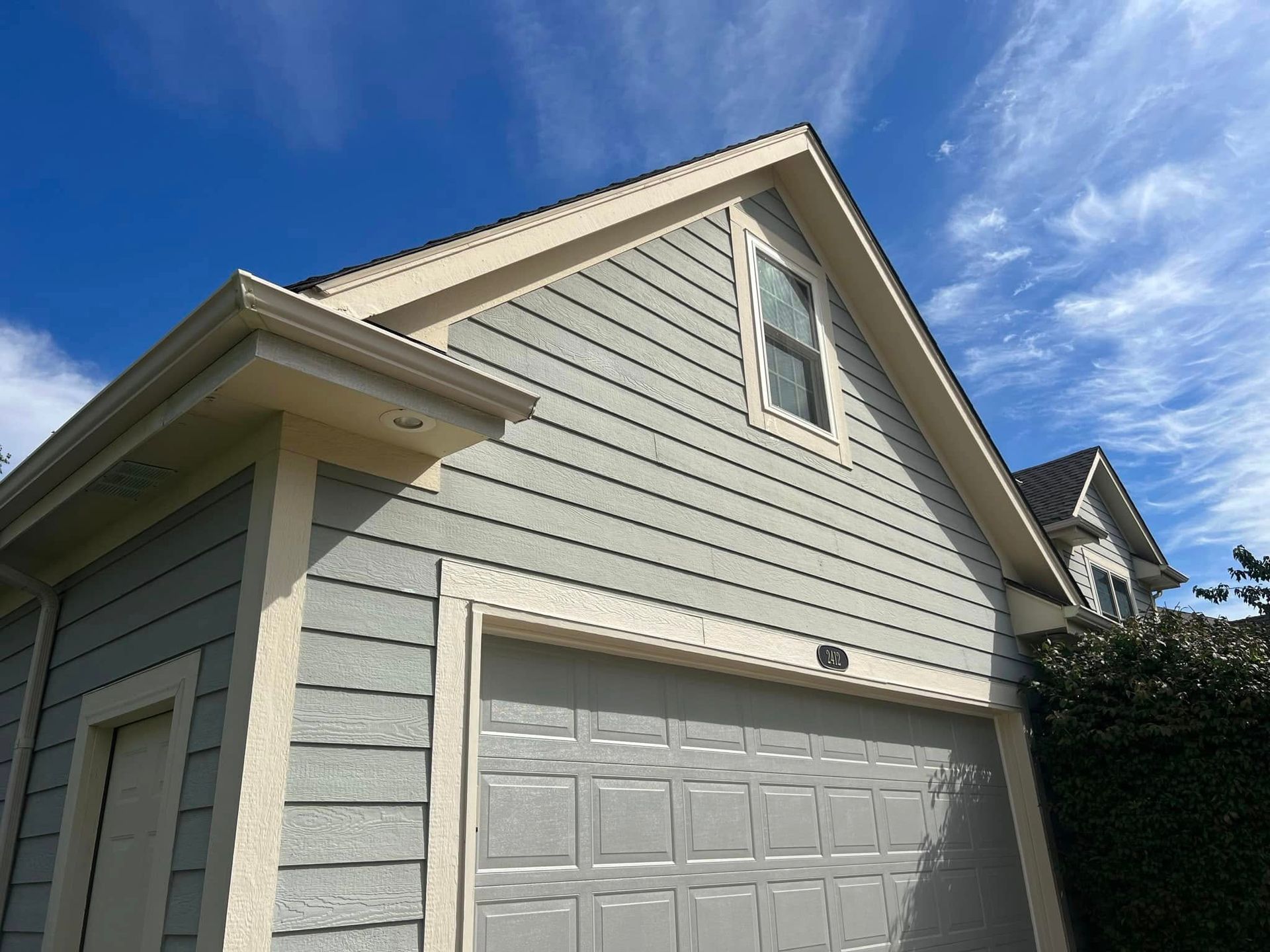 Garage with blue-gray siding, beige trim, a closed gray garage door, and a small window under a blue sky.