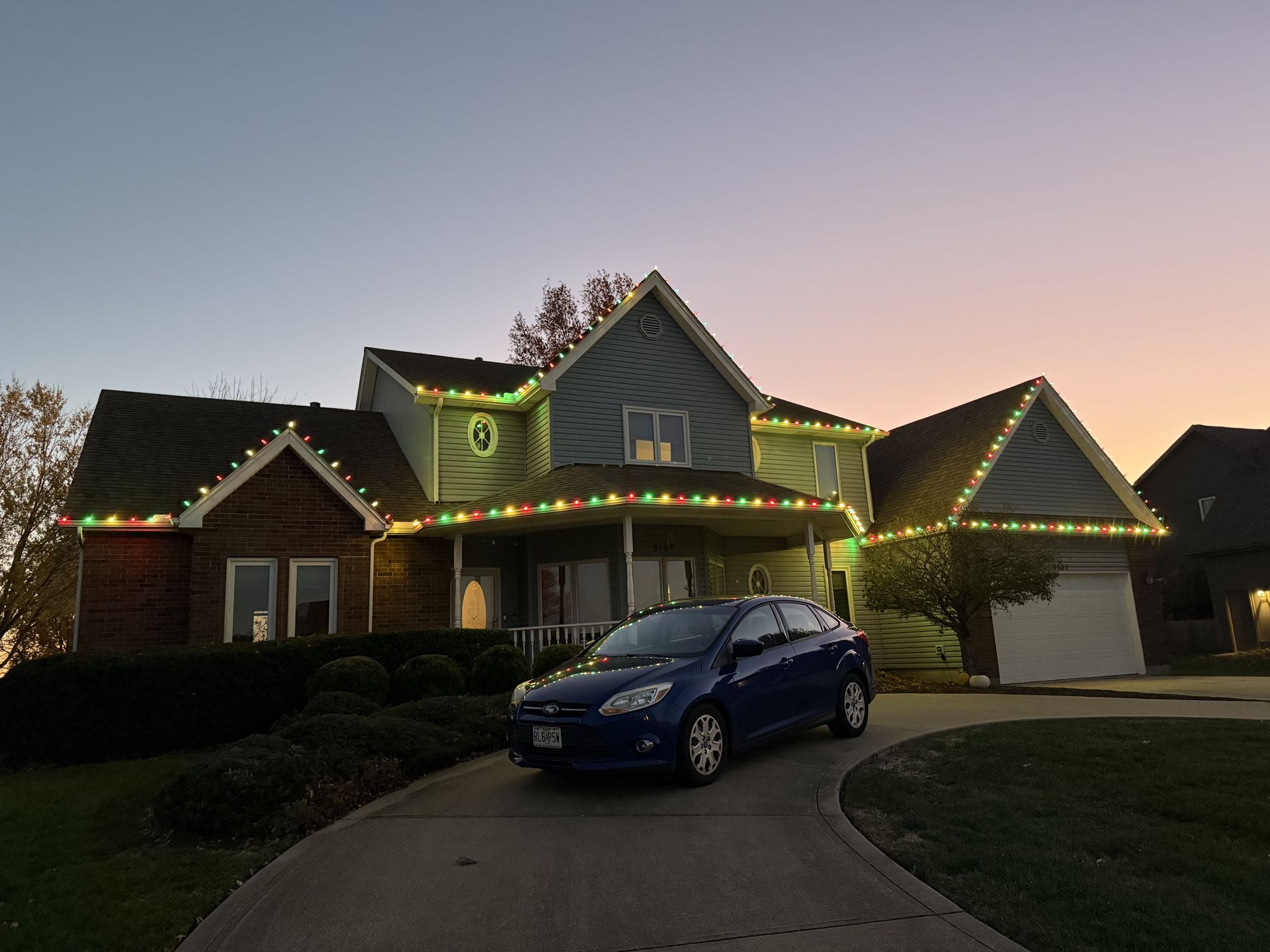 Blue car parked in driveway of house decorated with Christmas lights at dusk.
