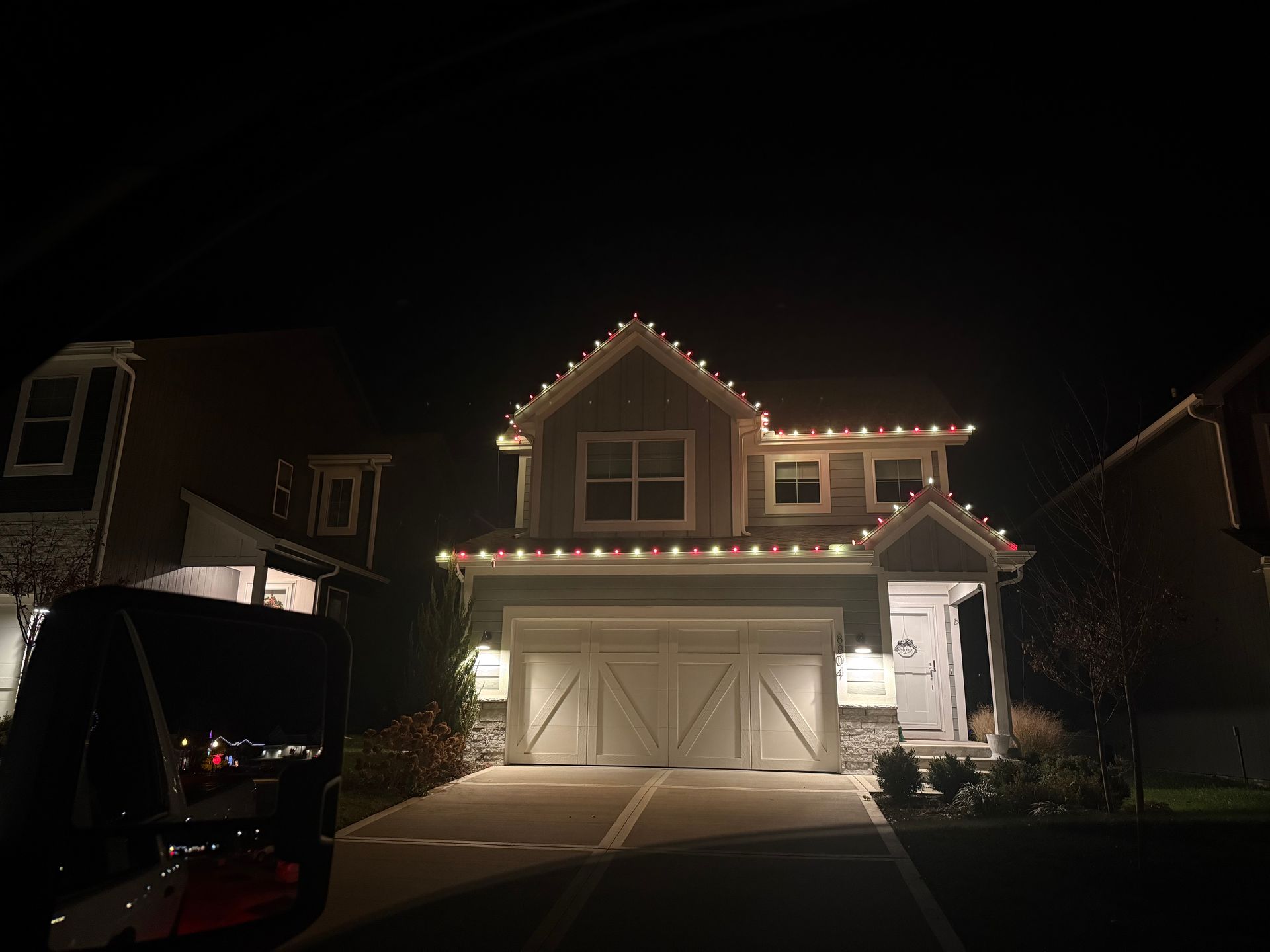 House at night decorated with red and white Christmas lights, lit garage and driveway.
