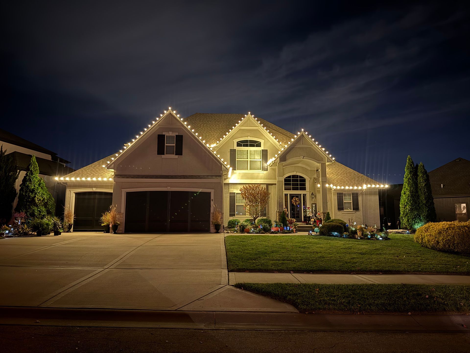 House at night, illuminated with white lights along rooflines, garage, and front yard landscaping.