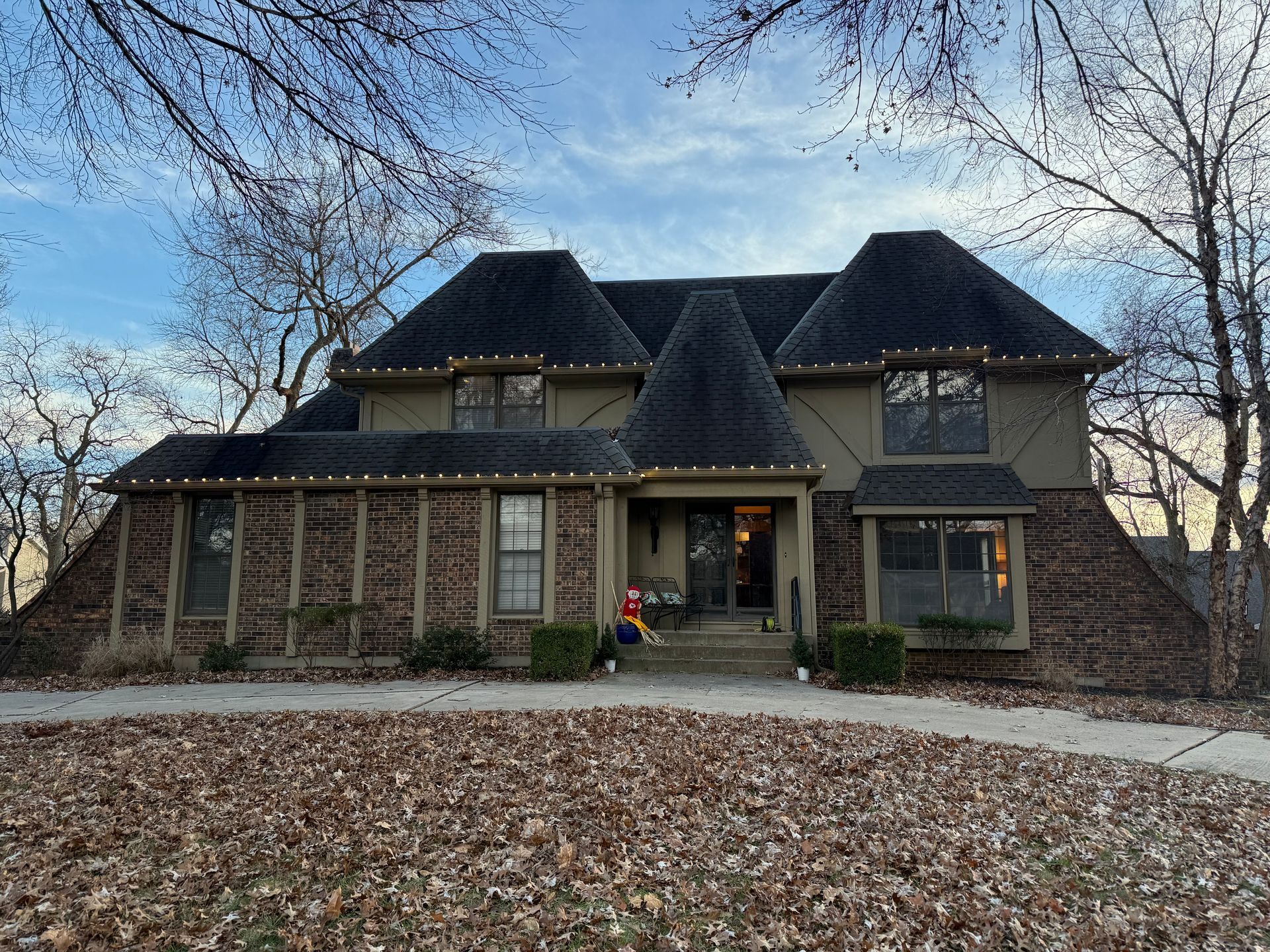 House with dark roof, brick facade, and Christmas lights. Brown leaves cover the yard.