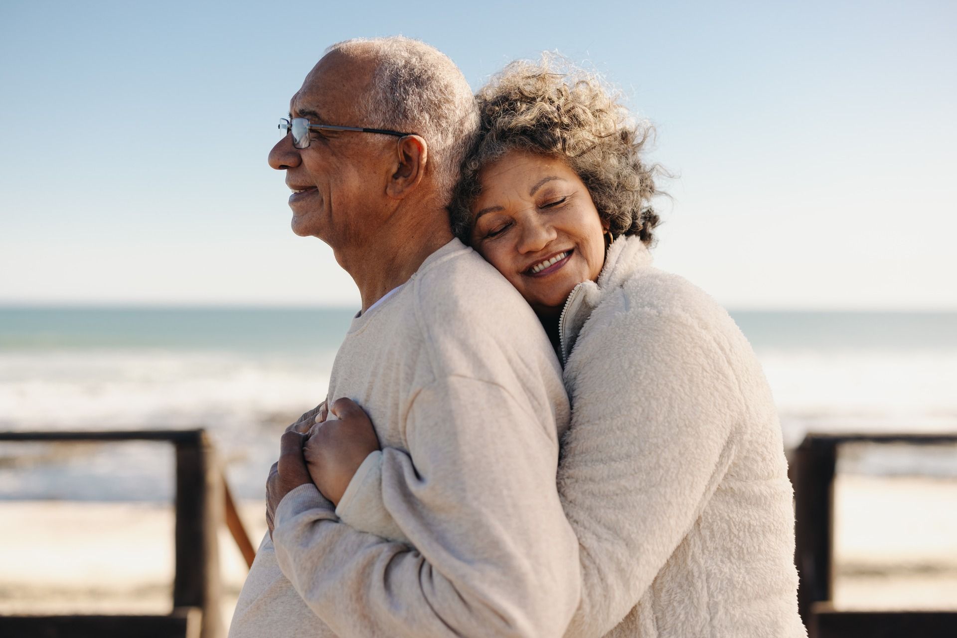Elderly couple embracing at the beach, woman smiles, sea and blue sky in the background.