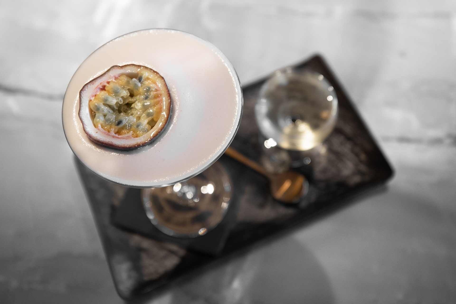 Een cocktail in een glas, gegarneerd met een halve passievrucht, op een dienblad met een lepel.