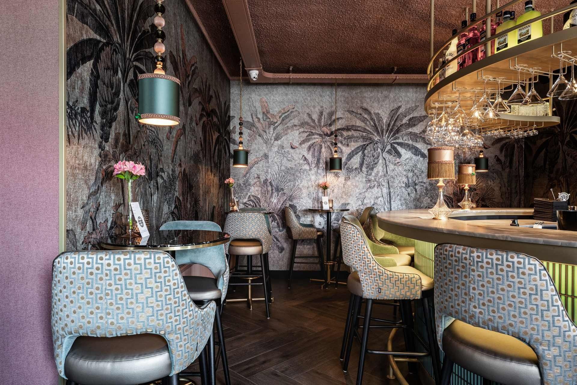 Barinterieur met behang met patroon, barkrukken en een ronde bar met een vitrine vol wijnglazen.