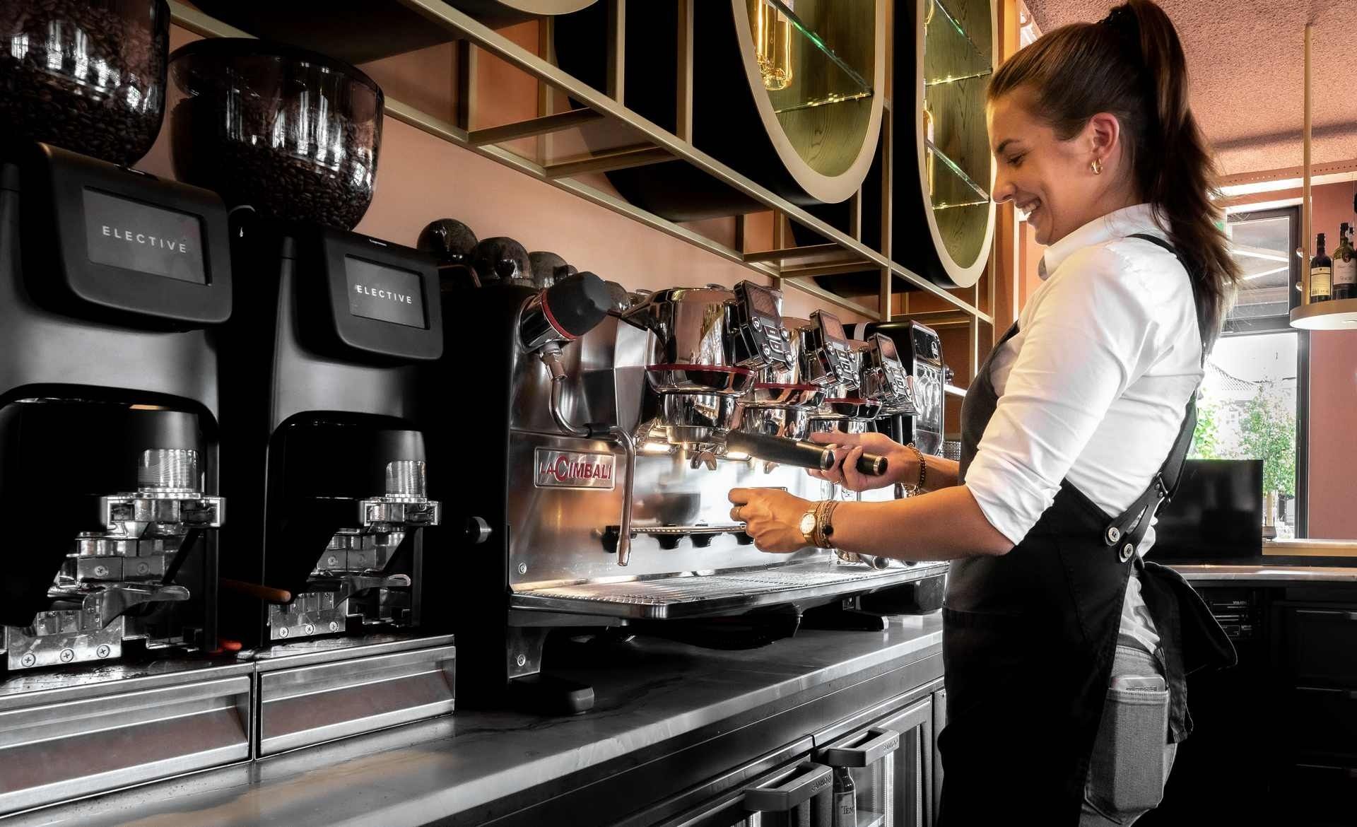 Een barista bedient met een glimlach een espressomachine in een café met roestvrijstalen apparatuur.