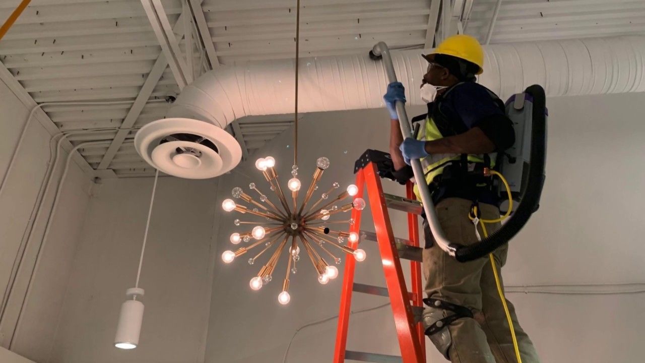 Man in safety gear cleaning air duct on a ladder in a white-walled room.