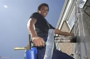 Man on ladder, smiling, cleaning a window. Blue sky and building exterior visible.