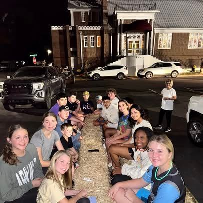 A group of young people are sitting on a hay bale in front of a building.