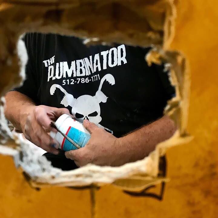 A person wearing a Plumbinator T-shirt holds a small container while viewed through a jagged hole in a yellow wall.