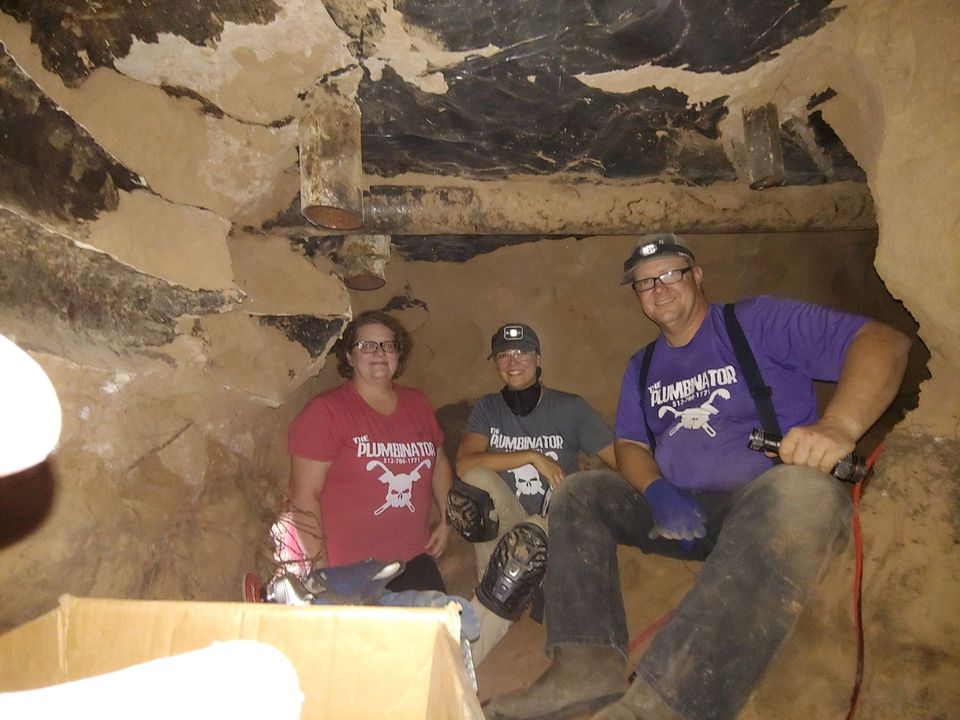 Three people wearing headlamps and shirts with a skull logo sit inside a dimly lit, earth-walled cave with wooden supports.