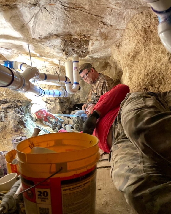 Two people working on plumbing pipes in a narrow, dirt-walled crawl space.