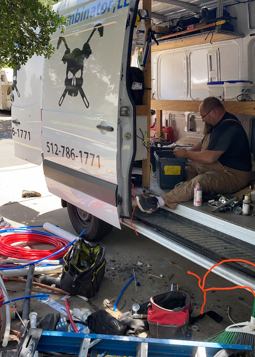 A worker sits in a van surrounded by plumbing tools and red and blue piping, with a logo showing a skull and wrenches.
