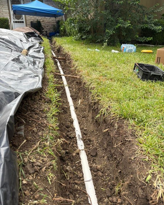 A white pipe lies inside a narrow trench dug into a green grass lawn next to a black plastic tarp.