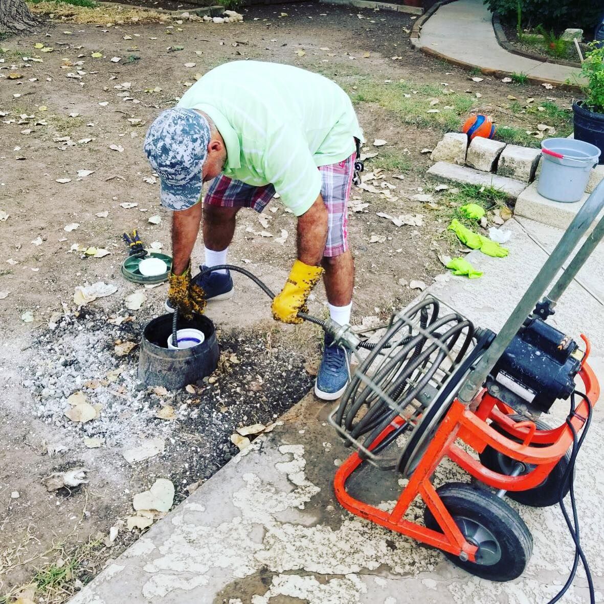 A person in a light green shirt and patterned shorts uses a sewer inspection camera to check an outdoor drain pipe.