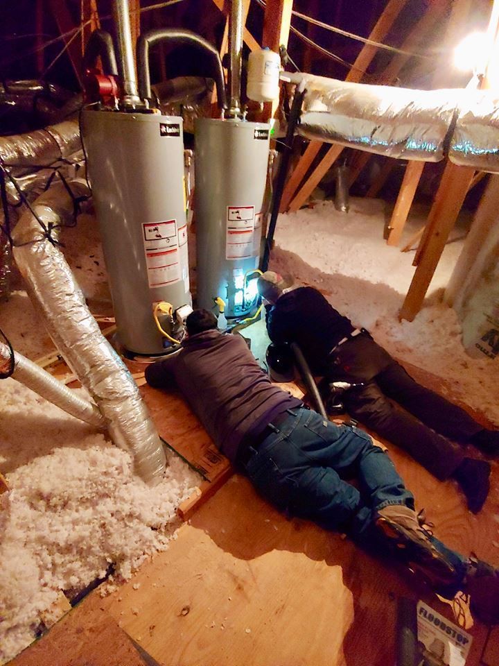 Two technicians work on water heaters in a dimly lit attic filled with white insulation.