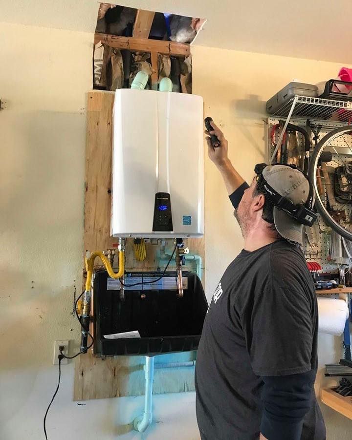 A technician examines a wall-mounted tankless water heater installed on a plywood board in a garage.