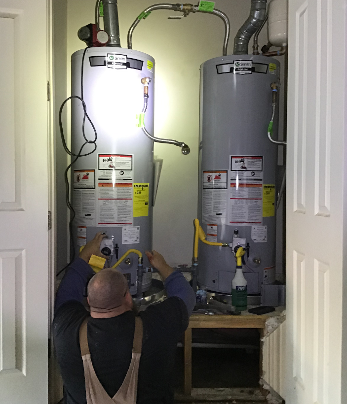 A worker in overalls kneels to repair two large grey water heaters inside a residential utility closet.