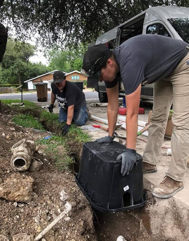 Two workers in workwear and gloves install a black plastic irrigation valve box in a trench beside a residential road.