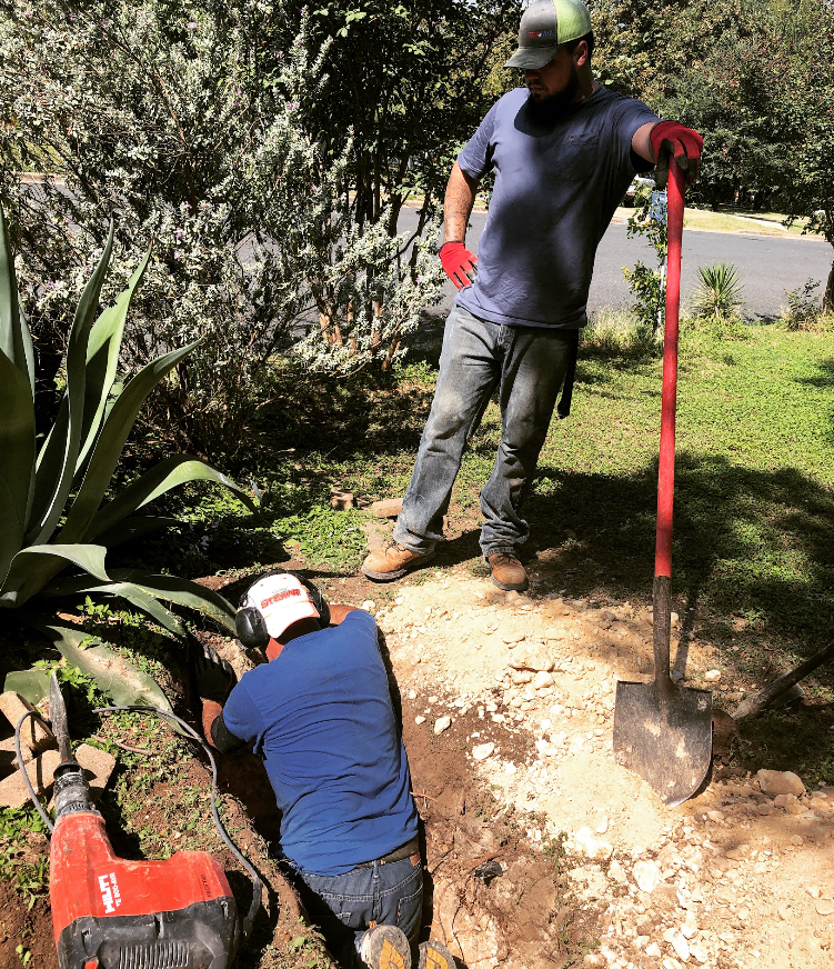 Two workers in a yard: one uses a red jackhammer in a trench, while the other stands nearby leaning on a shovel.