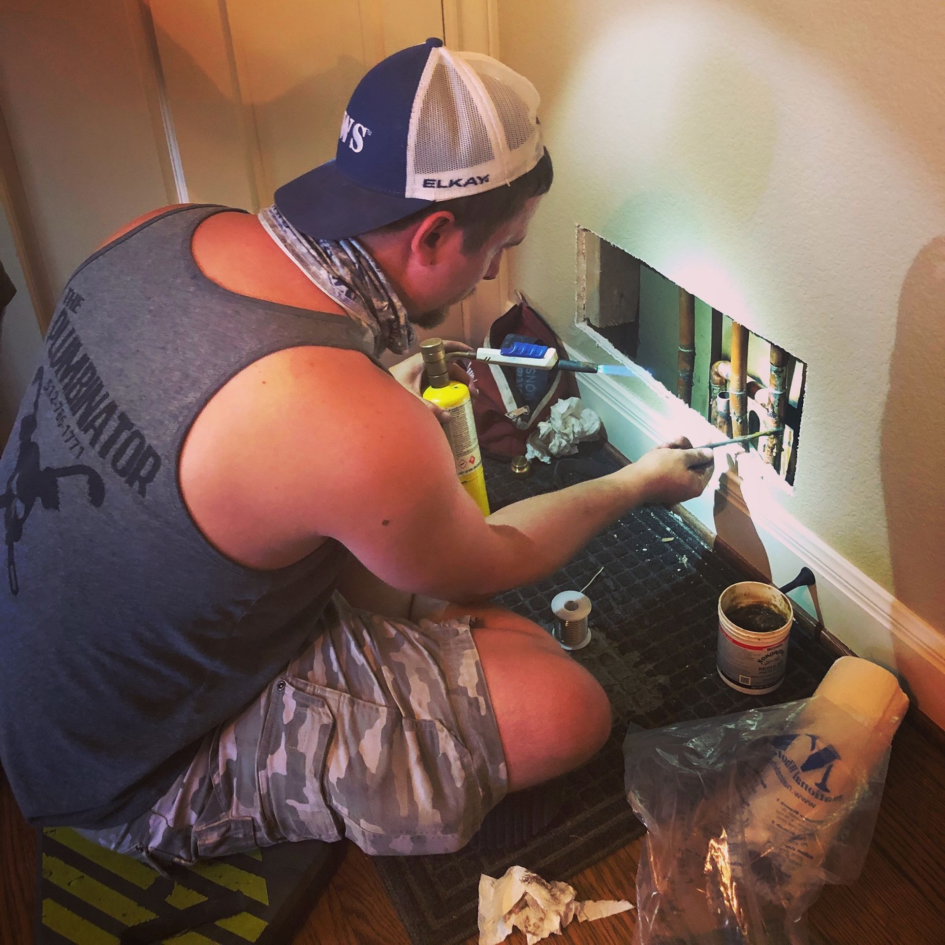 A person in a gray tank top and camouflage shorts uses a torch to solder copper pipes inside an open section of a wall.